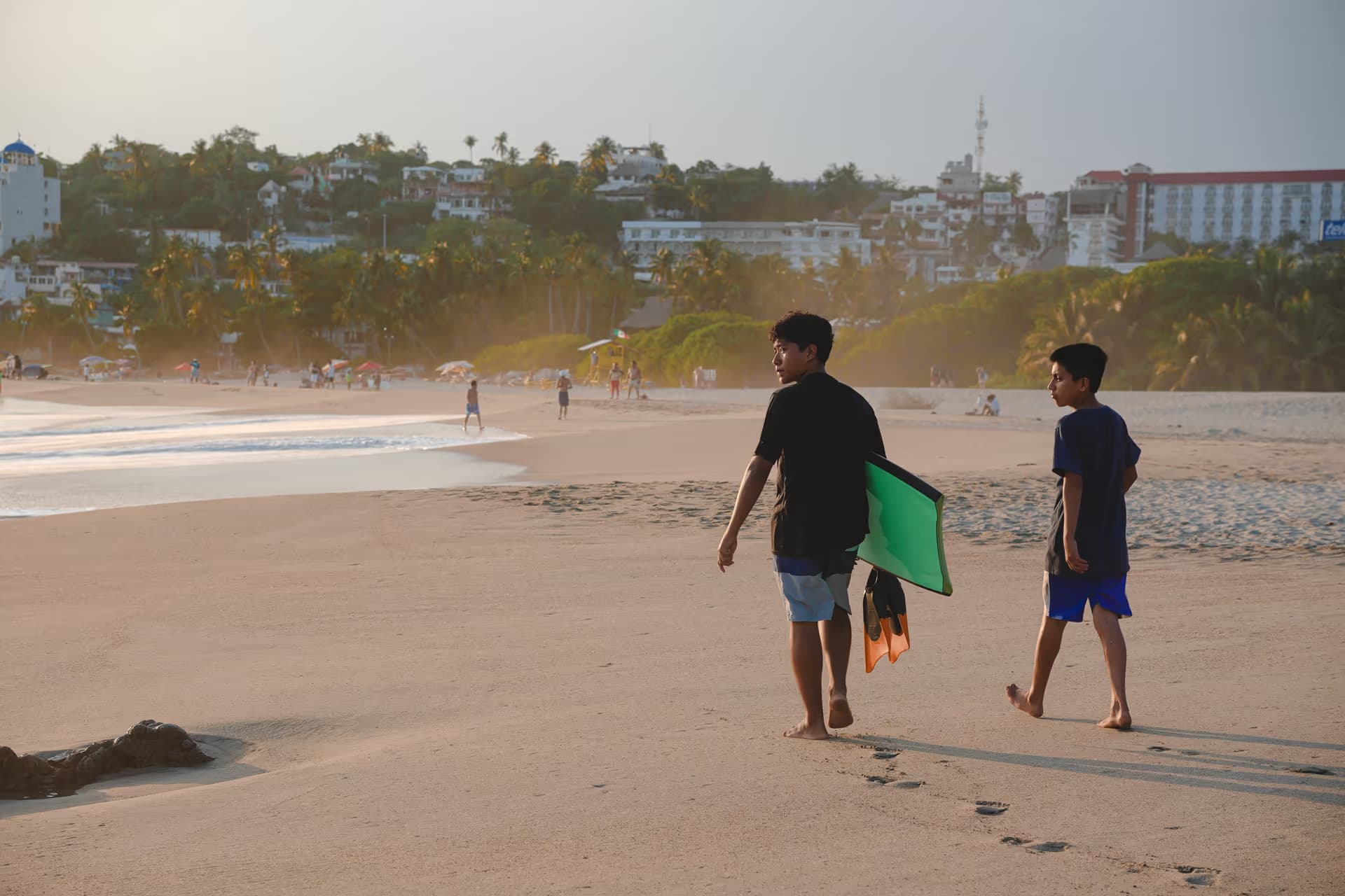 Dos personas caminando por la orilla al atardecer con una tabla de bodyboard